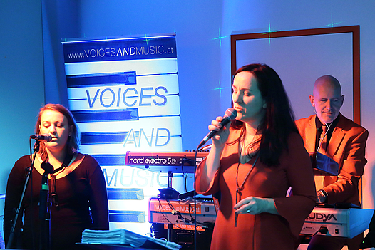 Zwei Sängerinnen im Vordergrund, ein Keyboardspieler und ein Roll-up von Voicesandmusic.at im Hintergrund