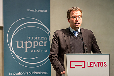 Ein Mann steht bei einem Rednerpult und spricht. Im Hintergrund ist ein Roll-up von Business Upper Austria zu sehen.