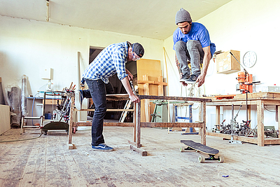 Ein Mann arbeitet auf einer Werkbank, zweiter Mann springt über die Werkbank und lässt sein Skateboard währenddessen unter der Werkbank durchfahren.