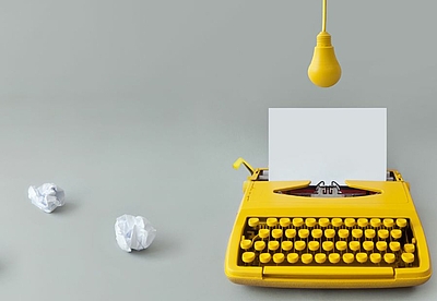 Gelbe Schreibmaschine mit eingespanntem Papier. Darüber eine gelbe Glühbirne. Daneben 3 zerknüllte weiße Papiere ©AdobeStock/Pixelbliss